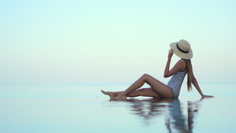 a sexy young woman in a bathing suit and straw sun hat sits on the infinity edge of a resort swimming pool