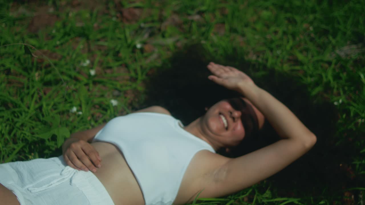 A woman dressed in white lies on green grass, shielding her face from sunlight with her hand
