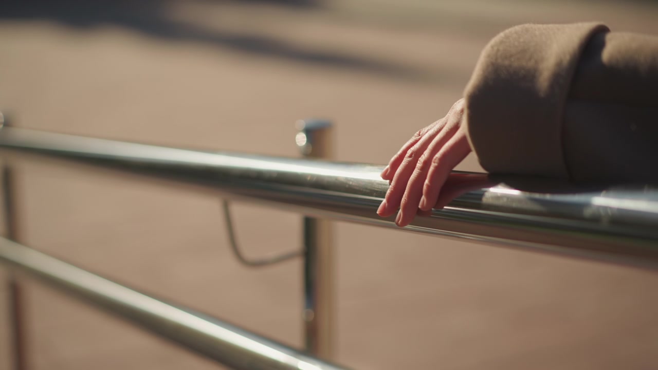 Close up of woman hand resting on polished metal railing with soft sunlight highlighting skin texture and fabric details. Background shows blurred outdoor pavement under warm daylight