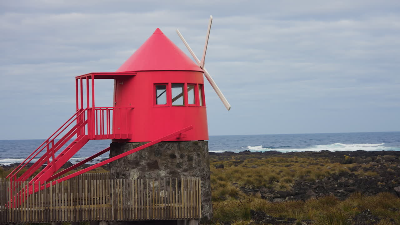 toma cinematográfica de un pequeño molino de viento rojo ubicado en la costa rocosa