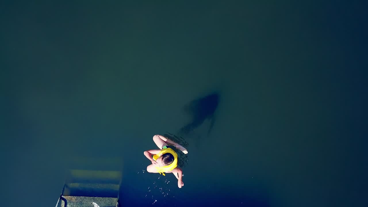 Close-up of a little kid jumping into the water from the old wooden bridge. Young boy in yellow vest dives into the river in summer. Motion camera bottom up