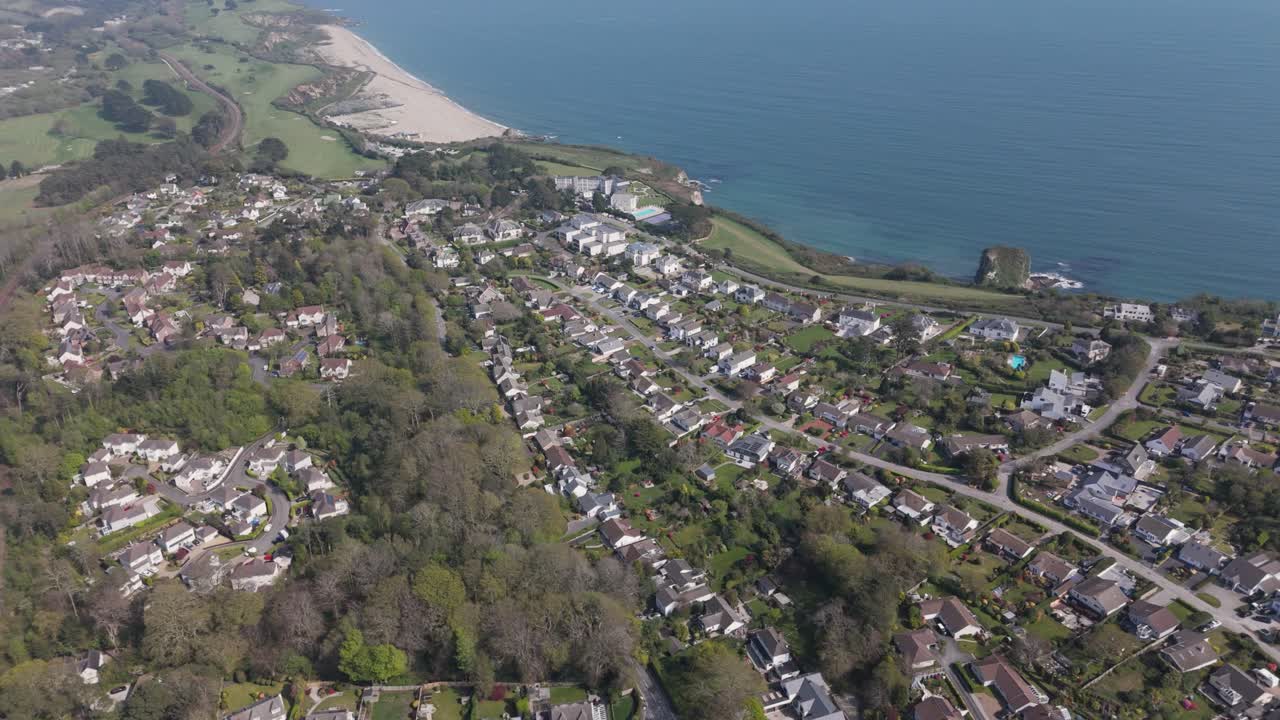 Aerial sweep over coastal suburb where curved streets of pitched-roof homes and gardens descend toward wide sandy beach and glittering sea below Cornish cliffs