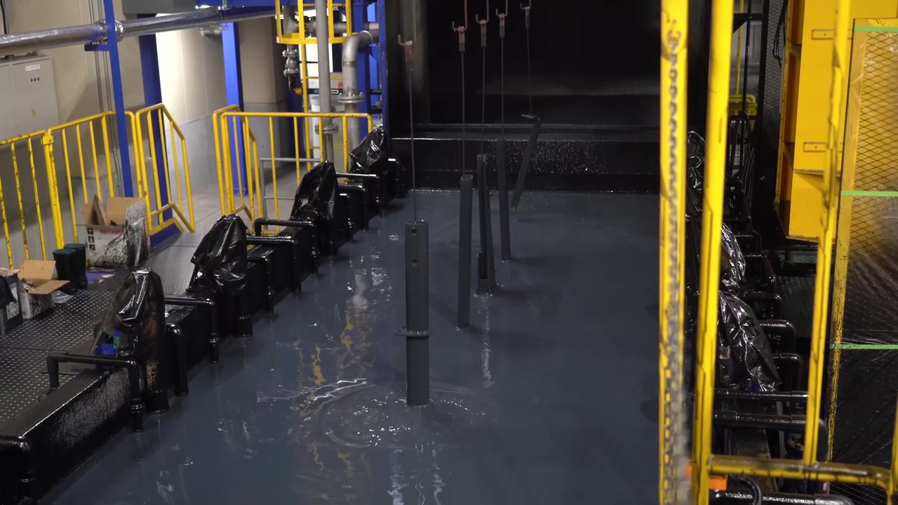 Overhead conveyor system carrying metal parts above a blue chemical washing tank behind yellow safety railings in a coating workshop
