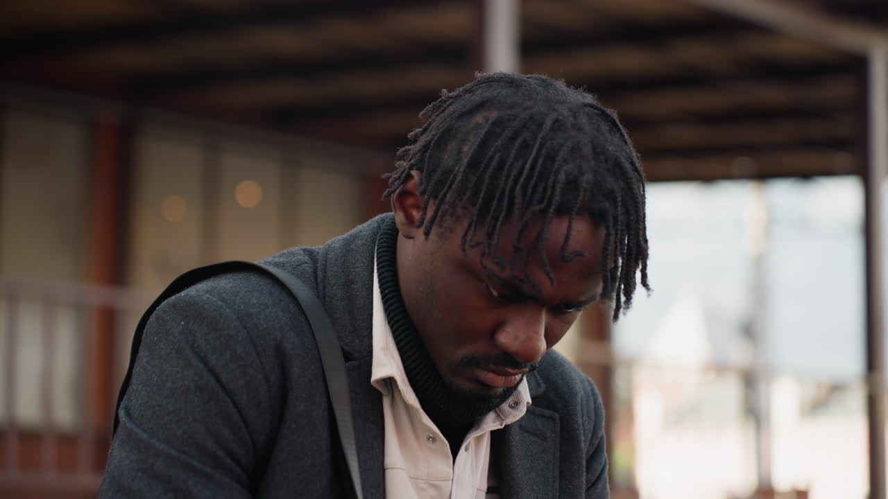 Close view of dreadlocked man with rough twisted natural hair looking down at mobile phone, dressed in layered clothing, showing style, and natural expression in soft daylight urban setting