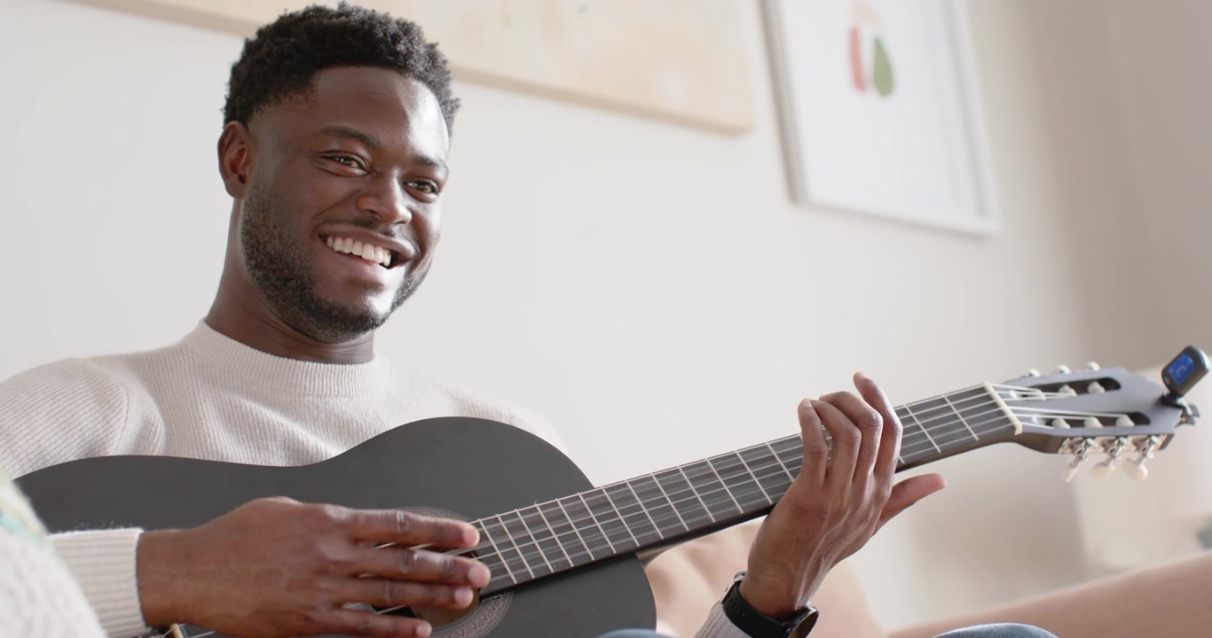 Uomo afroamericano sui 20 anni che ride, strimpella una chitarra nera a casa mentre si esercita su una canzone, con un maglione e un orologio.
