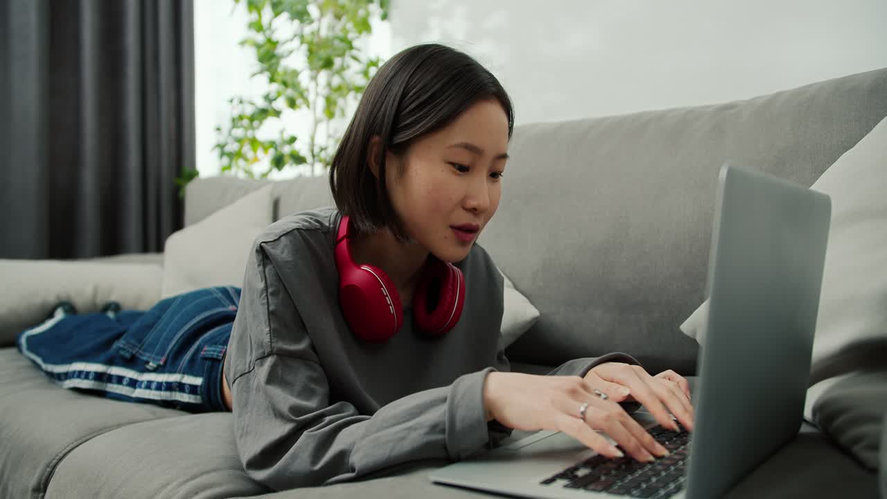 소파에 누워서 집에서 거실에서 일하는 아시아 여인이 노트북에서 타이핑하고 웃고 있습니다.