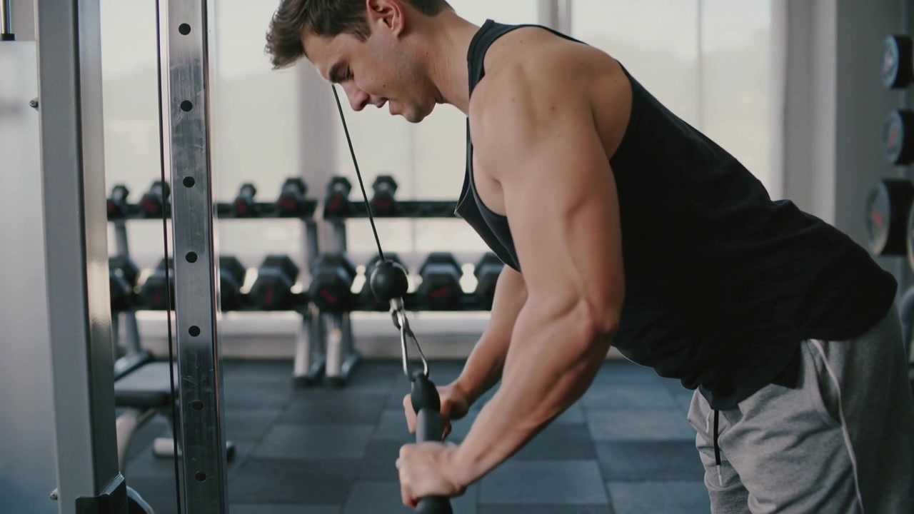 En mann i svart singlet utfører triceps pushdowns med kabel fra en startposisjon med bøyde albuer på et treningsstudio, trener triceps.