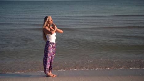 Cámara-Lenta-De-Mujer-Madura-En-Sarong-En-Una-Pose-De-Oración-En-Una-Playa-Al-Amanecer-O-Al-Atardecer