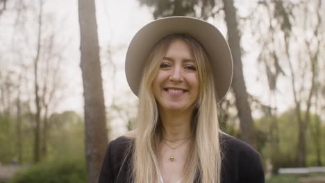 retrato de una mujer rubia feliz con sombrero de pie en el parque y mirando a la cámara