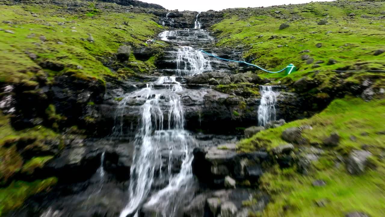 una suave cascada en la ladera de la colina fluye sobre escalones rocosos, rodeada de musgo verde y hierba