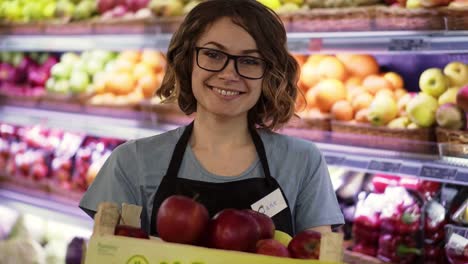 Schöne-Lächelnde-Junge-Weibliche-Supermarktangestellte-In-Schwarzer-Schürze,-Die-Eine-Kiste-Voller-äpfel-Vor-Dem-Regal-Im-Supermarkt-Hält,-Mit-Hübschem-Gesicht,-Das-In-Die-Kamera-Blickt-Professionelles-Frontporträt-Startgeschäft-1