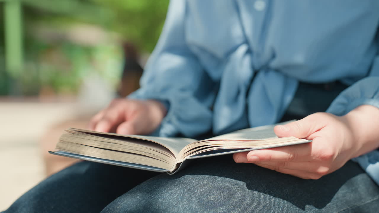 햇살이 비치는 공원에서 무릎에 소설책을 올려놓고 여유롭게 페이지를 넘기며 즐기는 차분한 백인 여성, 매듭이 있는 블라우스와 데님 스커트, 손과 종이를 클로즈업, 나뭇잎 보케와 함께 따뜻하고 편안한 분위기