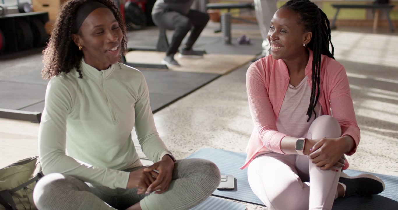 African American friends in workout clothes sitting on exercise mats at gym, chatting after rest