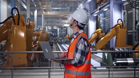 side view of asian male engineer with safety helmet working on a laptop and looking around while standing with industrial robotic arms working