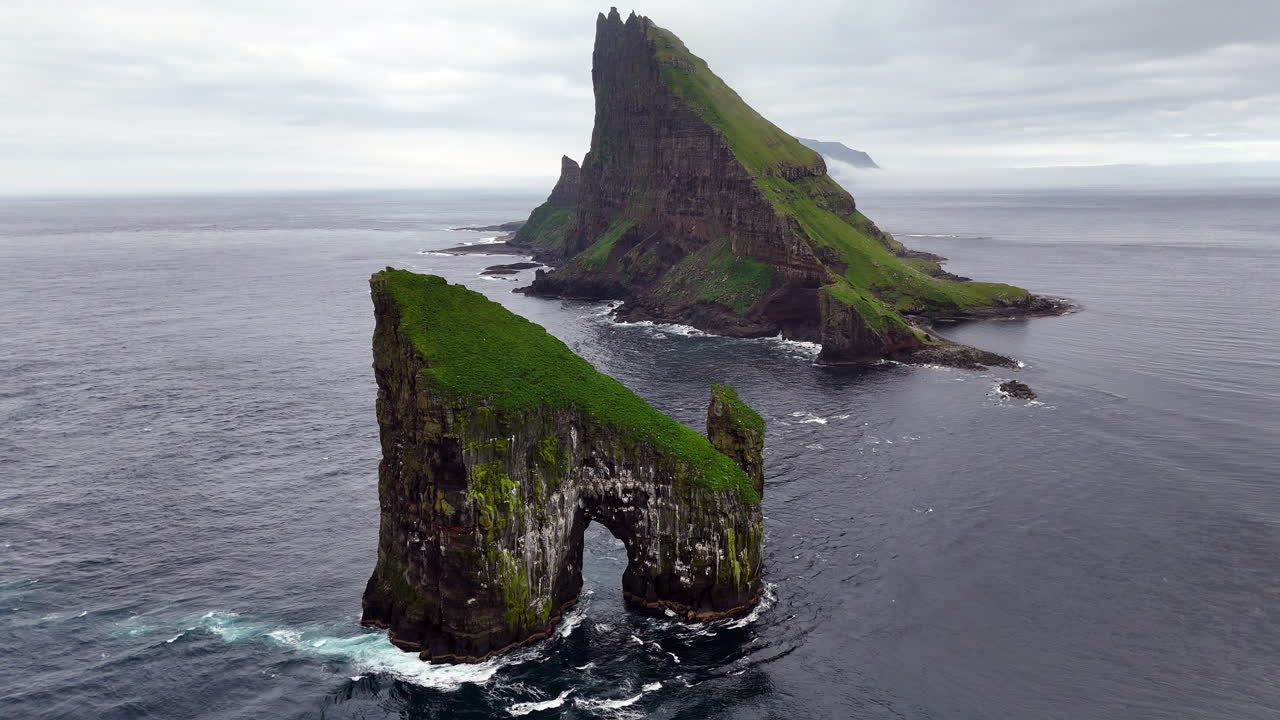 Cinematic aerial view of Drangarnir sea stacks rising dramatically from the Atlantic Ocean near Vágar, Faroe Islands, showcasing rugged cliffs, lush green slopes, and misty Nordic seascape
