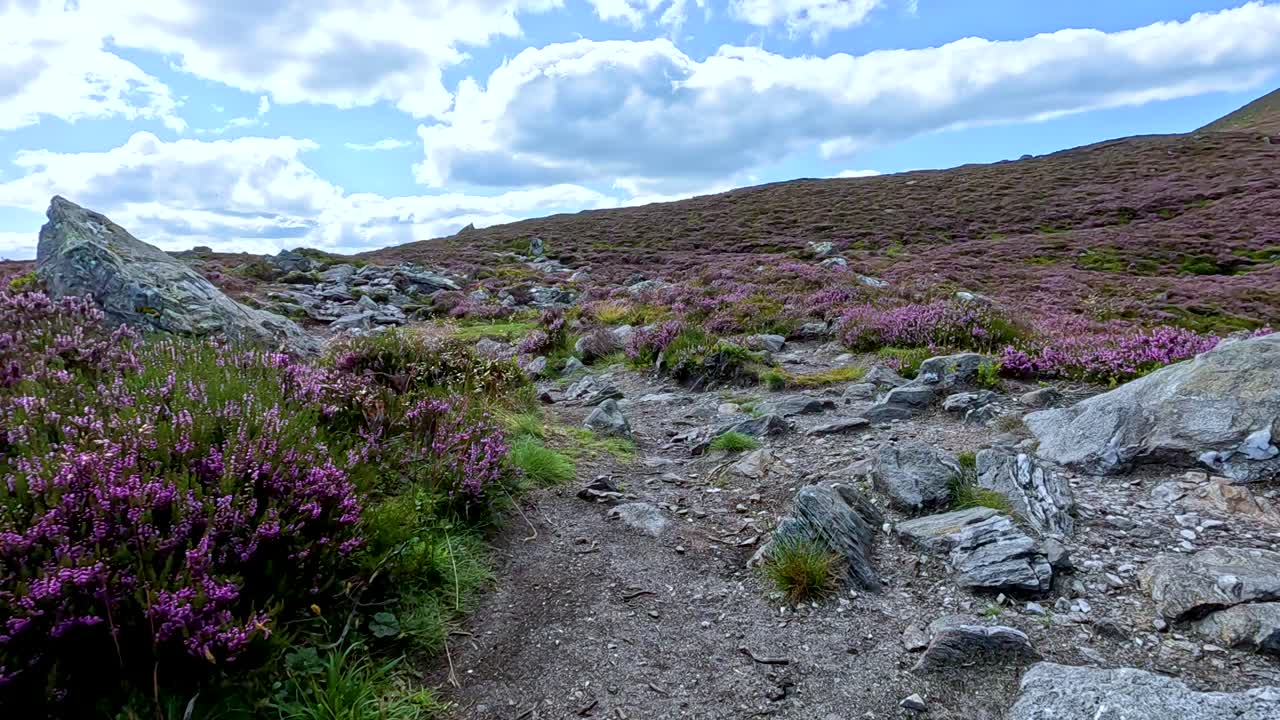A first-person view progresses along a rocky, heather-lined hiking trail under partly cloudy skies in the hills of Glen Clova, Scotland. Natural daylight, steady forward camera movement