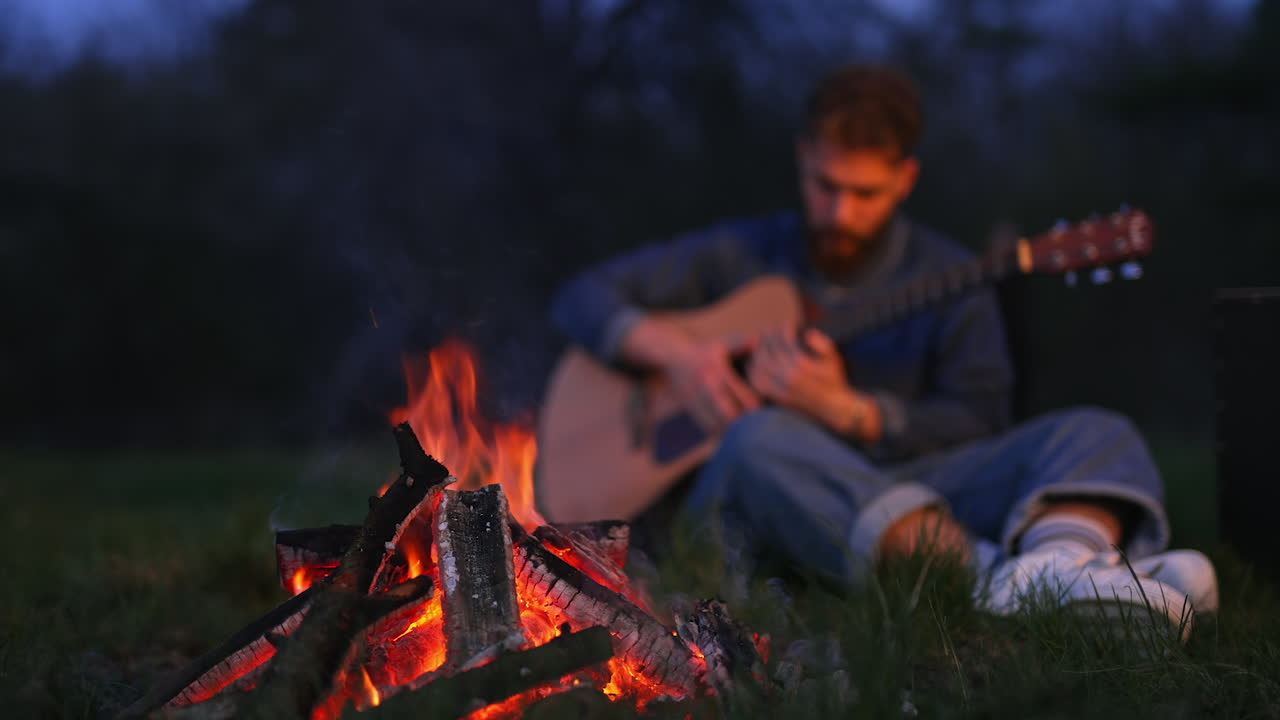 Bonfire burning at the campsite in the forest. Young man sitting by the fire and holding guitar in blur. Cozy summer evening with the company in the forest.