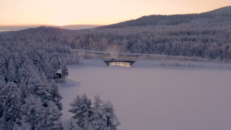 Aerial-view-tracking-vehicle-driving-across-misty-ice-lake-bridge-in-snow-covered-Scandinavian-woodland-hills