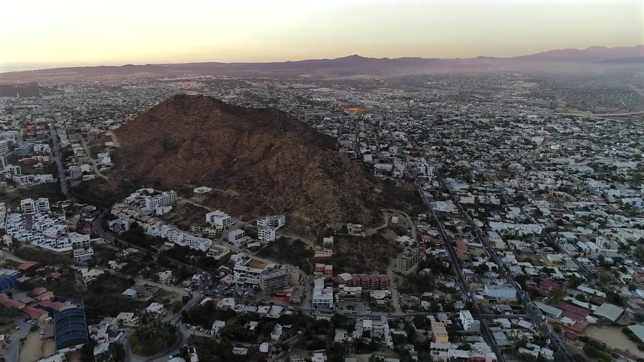 Drone rotates past the Pedregal mountains, revealing the northern cityscape of Cabo San Lucas across the land, with a distant mountain line visible far on the horizon beneath a fading sunset sky.