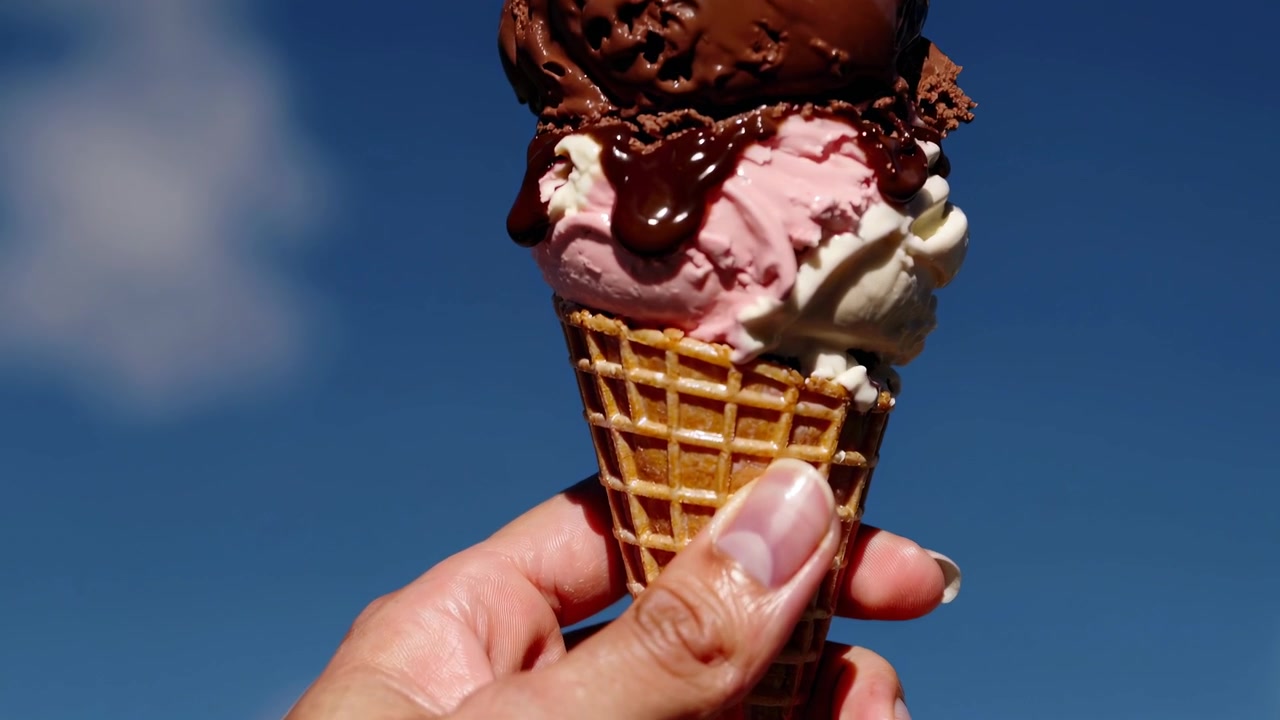 Mano che regge un cono gelato a più gusti con ciliegia contro il cielo blu