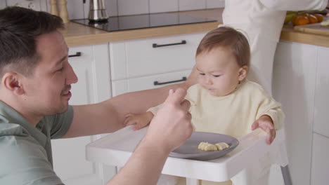 happy father feeding his cute baby girl sitting in her high chair in the kitchen while mother cutting fruit behind them 1