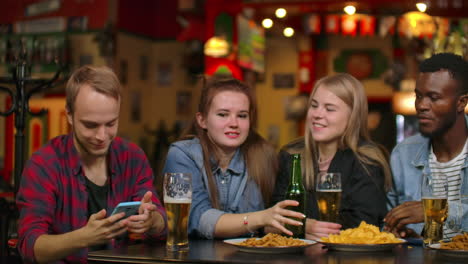 un hombre en compañía de amigos propone tomar una selfie grupal de una seda en un bar con una cerveza.