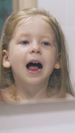 funny little girl with blue eyes looks at clean teeth and screams at large mirror in light bathroom at home close view