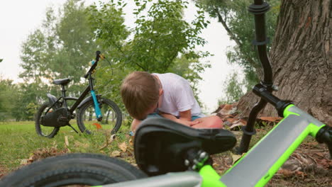 a child sits on a mat with his head bowed near a large tree trunk, a green bicycle lies on the ground in front of him while a black bicycle is parked behind