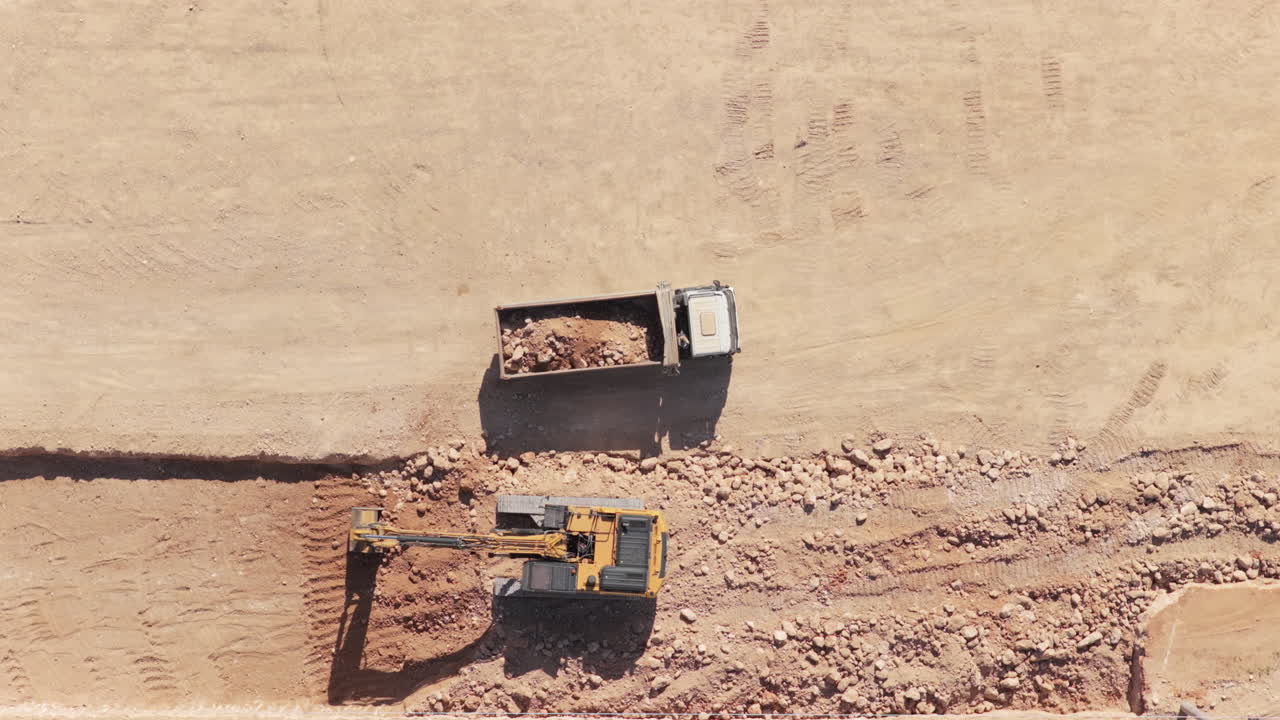Top-down aerial view of a yellow excavator digging a trench and loading the excavated soil and rocks into a dump truck, highlighting heavy machinery operations on a construction site