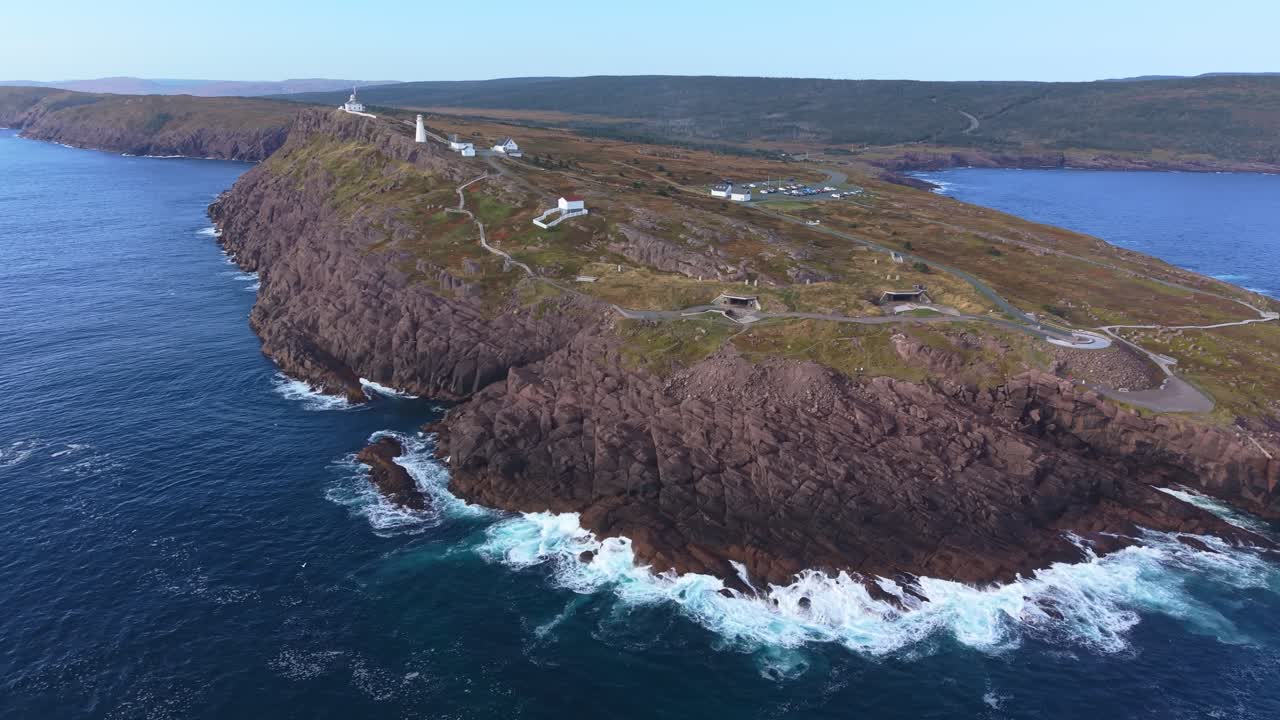 An elevated aerial captures Cape Spear's rugged coast with waves crashing against cliffs and Signal Hill visible far away, filmed from outside the national park boundary