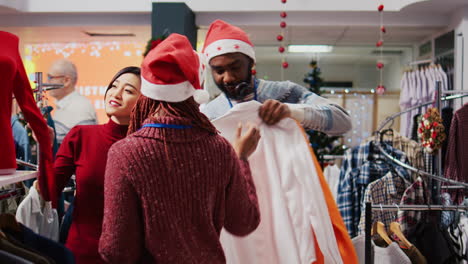 Friendly-retail-assistant-in-festive-ornate-shopping-mall-fashion-boutique,-showing-client-beautiful-red-garments-while-african-american-coworker-brings-stylish-blazers-for-her-to-try-on