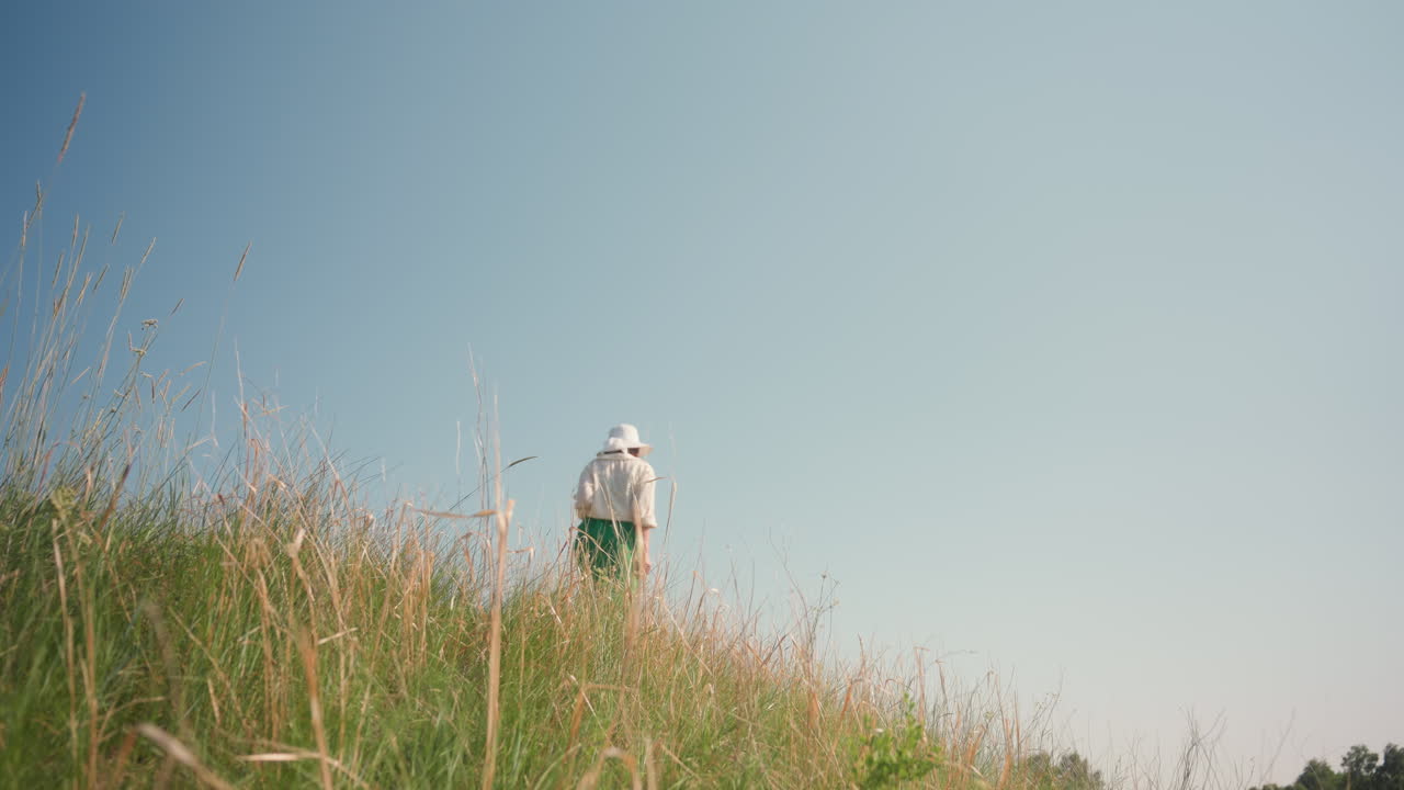 Young woman in white sunhat and green dress walks along grassy hilltop field edge, camera swinging by her side under bright blue sky, tall grass framing rural landscape while she explores outdoors