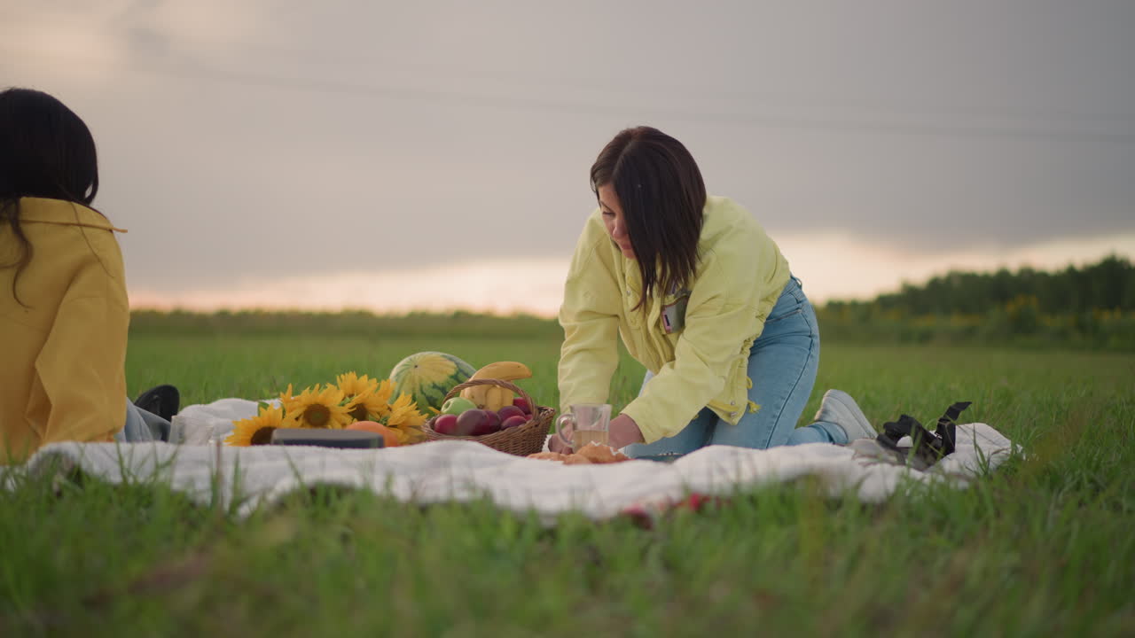 두 여성이 담요 위에서 피크닉을 즐기고, 초원 위로 해가 지는 석양, 캐주얼한 노란색 재킷, 해바라기 꽃다발과 과일, 편안한 대화와 웃음, 부드러운 황금빛, 멀리 보이는 넓은 잔디밭