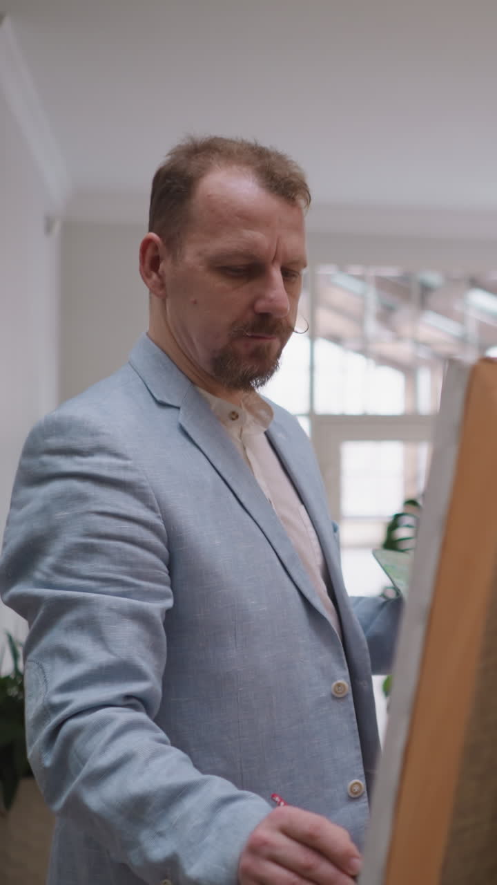 el artista trabaja en el caballete en el estudio. el diseñador pensativo dibuja obras de arte en grandes lienzos en el taller de arte. el hombre barbudo en chaqueta aplica color a la obra maestra