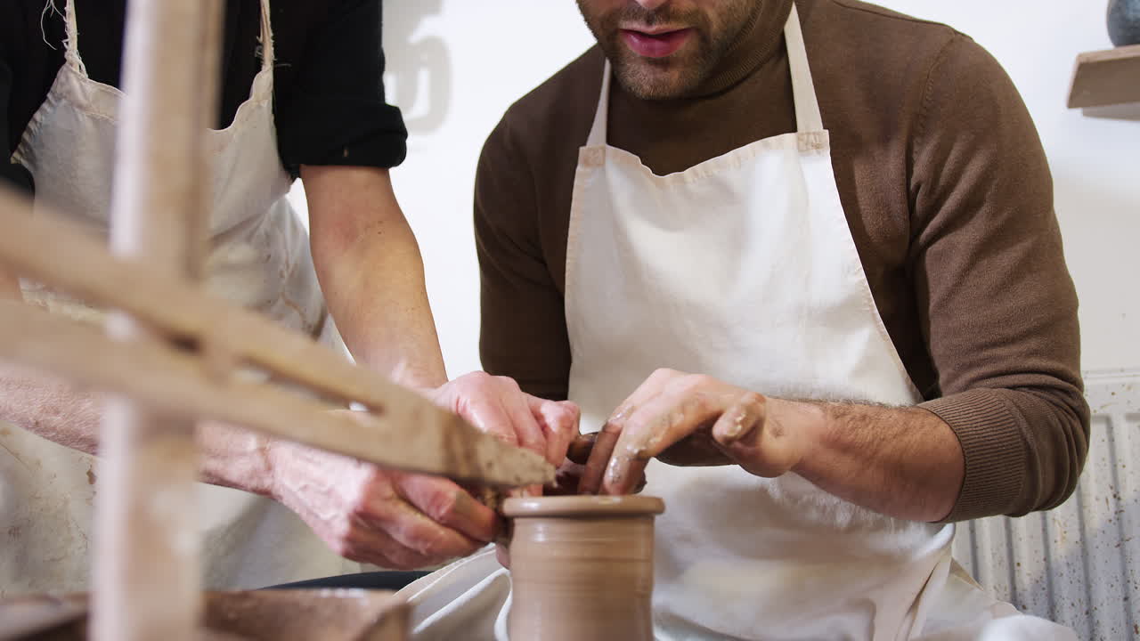 Close Up Of Male Teacher Helping Man Sitting At Wheel In Pottery Class