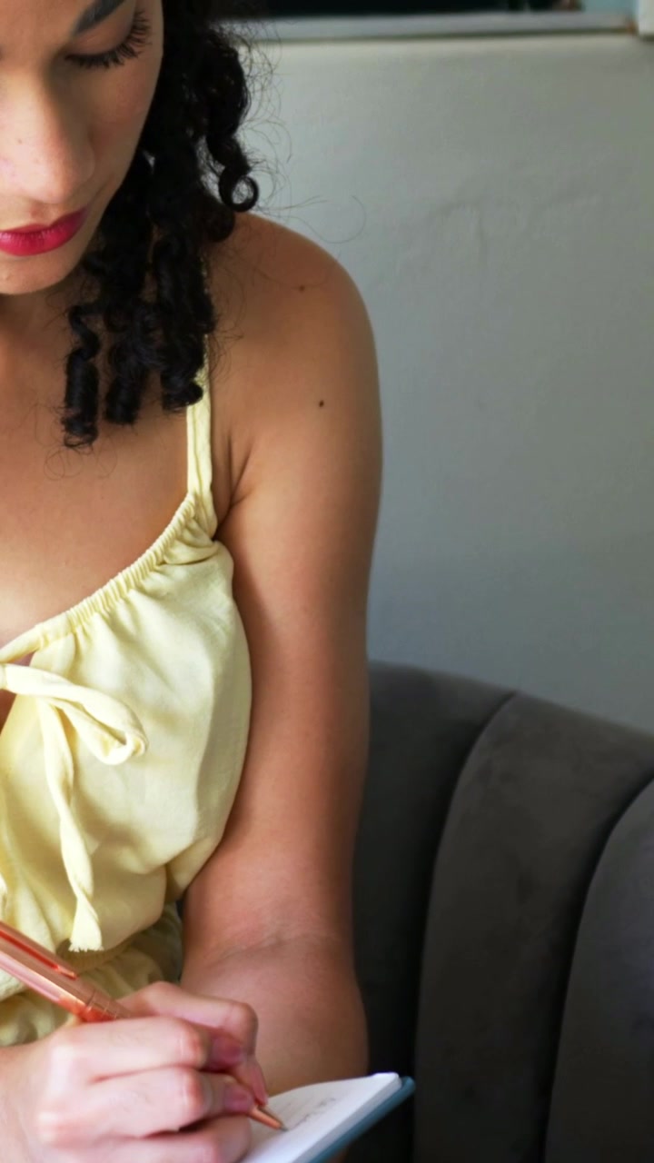 Video verticale: Zoom su una donna che scrive un diario su un quaderno sul divano con una penna, indossa un top giallo pallido, con spazio per il testo.