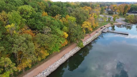 Gente-Caminando-Por-Un-Sendero-Alrededor-De-Un-Lago-Durante-El-Otoño-Junto-A-Un-Bosque-Con-Hojas-Coloridas-En-Los-árboles,-Muelle-De-Pesca-En-El-Fondo