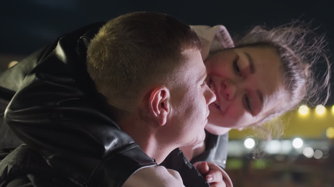 Close up of focused man working outdoors at night as woman holds him gently from behind while wind blows her hair under glowing city lights and soft night breeze in urban environment