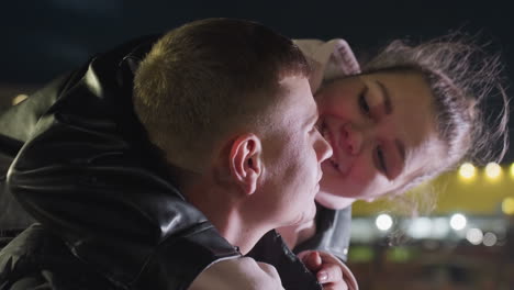 close up of focused man working outdoors at night as woman holds him gently from behind while wind blows her hair under glowing city lights and soft night breeze in urban environment