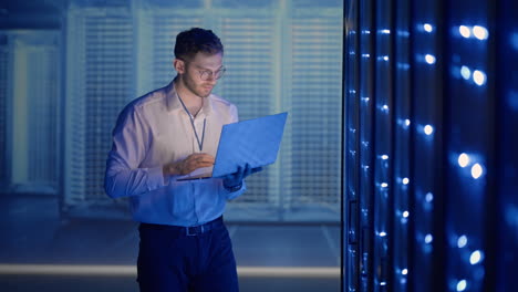 Male-network-engineer-doing-a-system-check-standing-in-the-server-room-with-his-laptop.-At-data-center-men-server-specialists-inspecting-working-system-and-hardware-of-rack-server-computer-cabinets