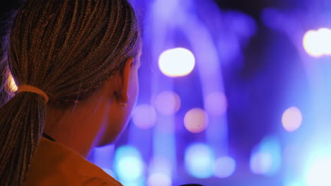 The-child-admires-the-fountain-with-lighting,-sits-on-the-edge-of-the-fountain,-rear-view