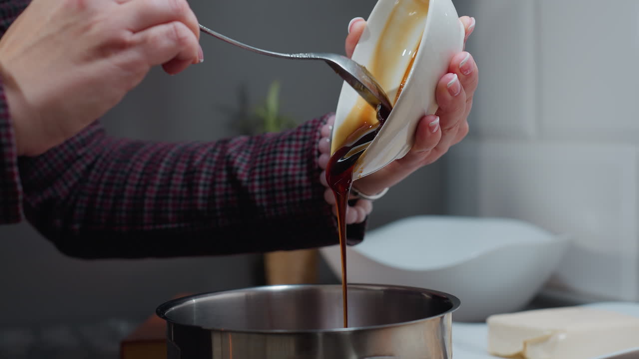 primer plano de la mano en la manga a cuadros vertiendo miel gruesa de un cuenco pequeño en la olla, un ambiente de cocina cálido con electrodomésticos borrosos, encimera e ingredientes en el fondo