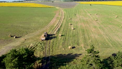 Tractor-Con-Remolque-Recogiendo-Fardos-De-Heno-Redondos-En-Tierras-Agrícolas,-Revelación-De-Drones