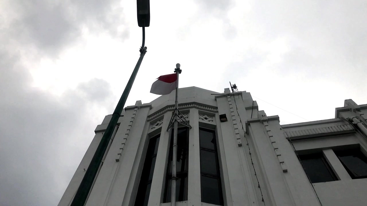 Ripresa 4K a mano dal basso della bandiera indonesiana che sventola in cima a un edificio coloniale bianco contro un cielo nuvoloso. Filmato patriottico del patrimonio storico.