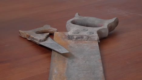 Zoom out revealing a rusty antique hand saw with a worn handle placed on a wooden table