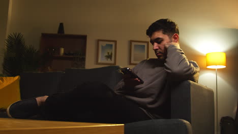 young man spending evening at home sitting on sofa with mobile phone scrolling through internet or social media 8