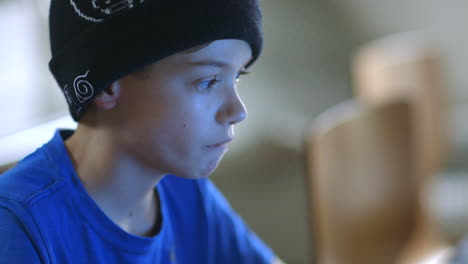 young boy wearing a sock hat nervously chews his lip while playing on a laptop