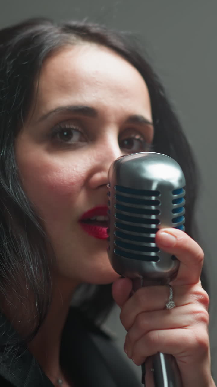 Close-up of a female vocalist in a black outfit, holding a vintage mic with one hand, singing with a serious expression as the camera zooms in and out against a blurry background