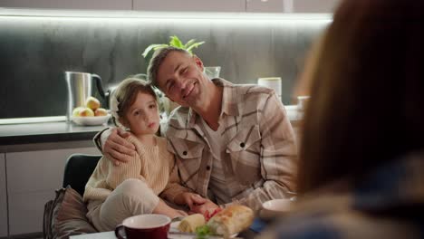 Over-the-shoulder-portrait-of-a-happy-middle-aged-man-with-gray-hair-along-with-his-little-daughter-with-brown-hair-with-a-braided-hairstyle-who-are-sitting-at-the-table-and-chatting-together-with-a-brunette-woman-during-a-family-dinner-in-the-evening