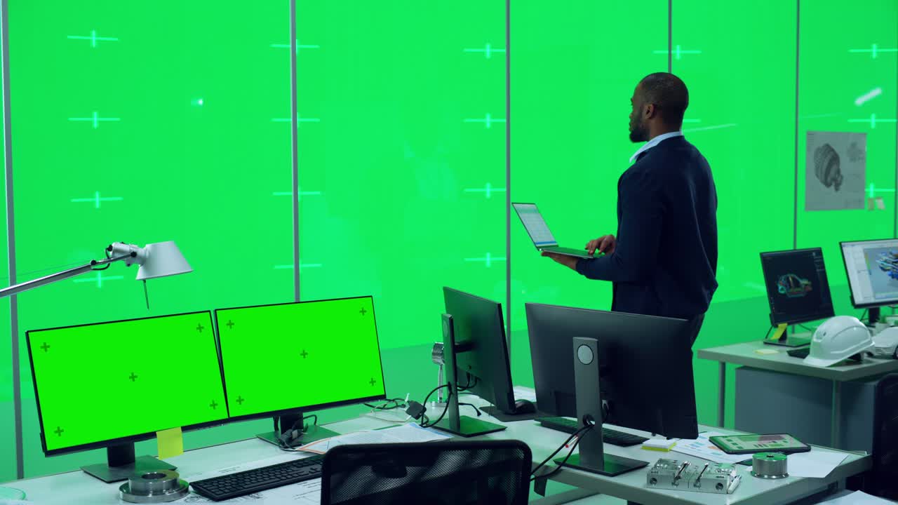 un bell'ingegnere maschio nero in piedi in un ufficio con computer desktop di fronte a uno schermo virtuale verde chroma key wall con segnaposti per mock-up e modelli. modello utilizzando tablet.
